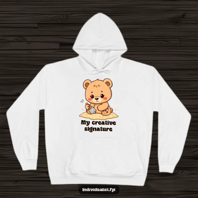 Funny, determined bear character diligently creating a unique pattern in the sand, depicted on a warm and cozy hoodie.